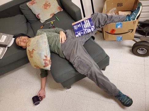 Student laying across a couch with pillow and book on top of him